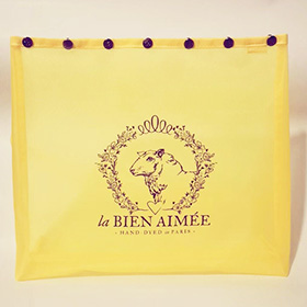 A yellow mesh bag with snaps on top and a photo of a sheep with a wreath around it and text that say la Bien Aimee