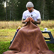 A model sitting on a bench crocheting a brown blanket